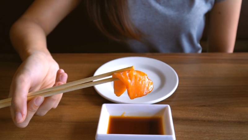 A person holding a piece of salmon sashimi with chopsticks over soy sauce on a wooden table