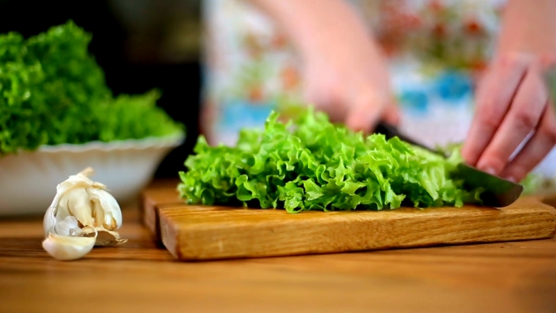 Fresh green lettuce being chopped on a wooden cutting board in a kitchen, with a clove of garlic placed nearby